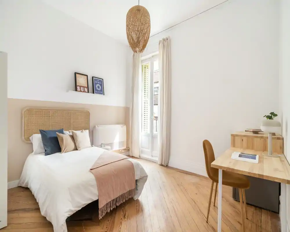 Chambre lumineuse et aérée avec tête de lit tressée, coussins superposés et plaid doux ; une grande fenêtre inonde la pièce de lumière naturelle et un petit bureau offre un espace de travail fonctionnel.