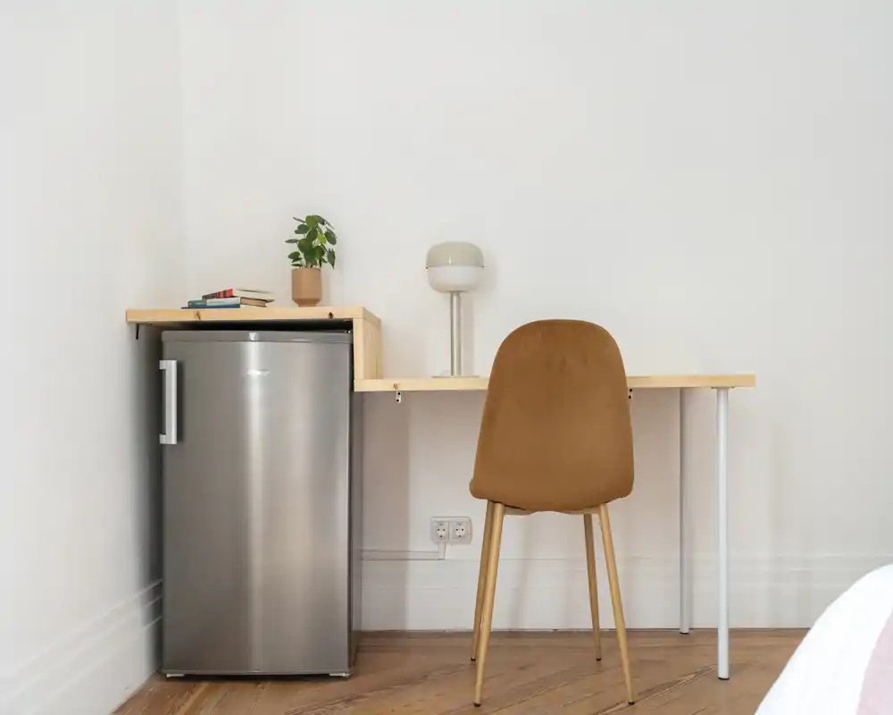 Coin de chambre simple avec un petit bureau, une chaise et un mini-frigo. Style épuré et minimaliste, une lampe et une petite plante apportent une touche chaleureuse.