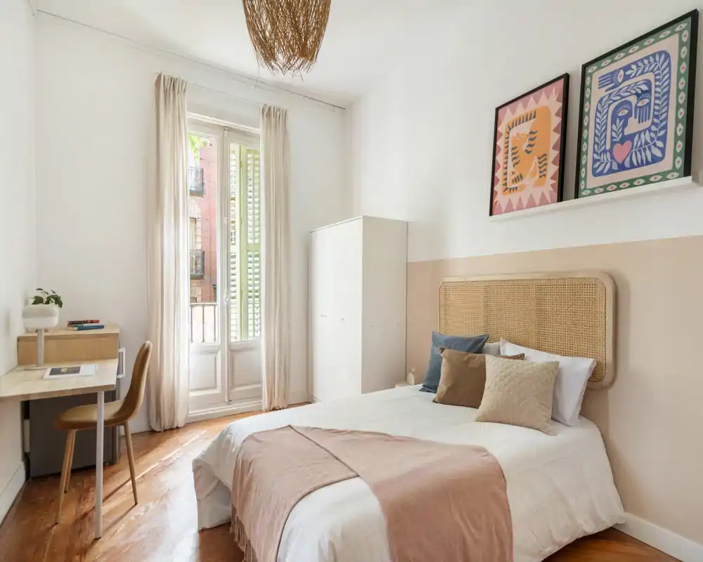 Chambre lumineuse et stylée avec tête de lit en rotin, coussins superposés et plaid rose; grande fenêtre aux voilages qui inonde la pièce de lumière naturelle et petit bureau pratique.