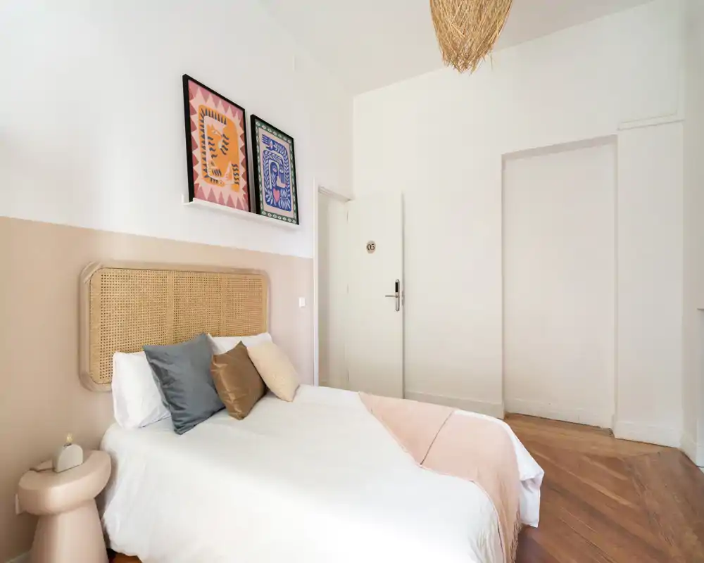 Chambre lumineuse et bien décorée avec une tête de lit en cannage, coussins superposés et accents pastel; parquet et lumière naturelle pour une ambiance chaleureuse.