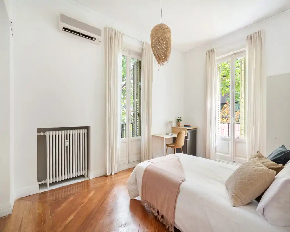 Chambre lumineuse et aérée avec de grandes fenêtres, parquet en bois et lit bien fait agrémenté de textiles doux et d'un luminaire en osier — idéale pour un séjour reposant.
