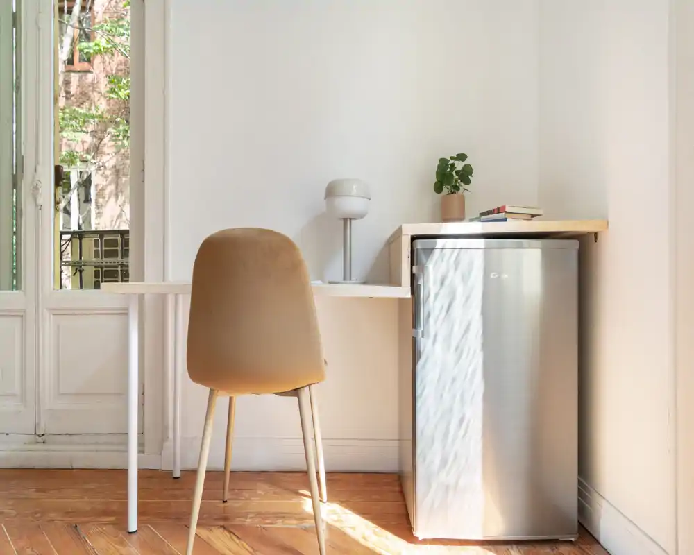 Coin kitchenette compact avec petit réfrigérateur en inox, plan de travail, lampe et chaise — lumineux, minimaliste et adapté à une annonce de studio.