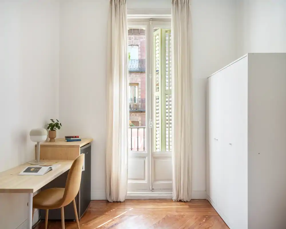 Chambre lumineuse et minimaliste avec grande fenêtre à guillotine et rideaux clairs, petit bureau et chaise, et une armoire blanche — parfaite pour une personne ou un espace de télétravail.