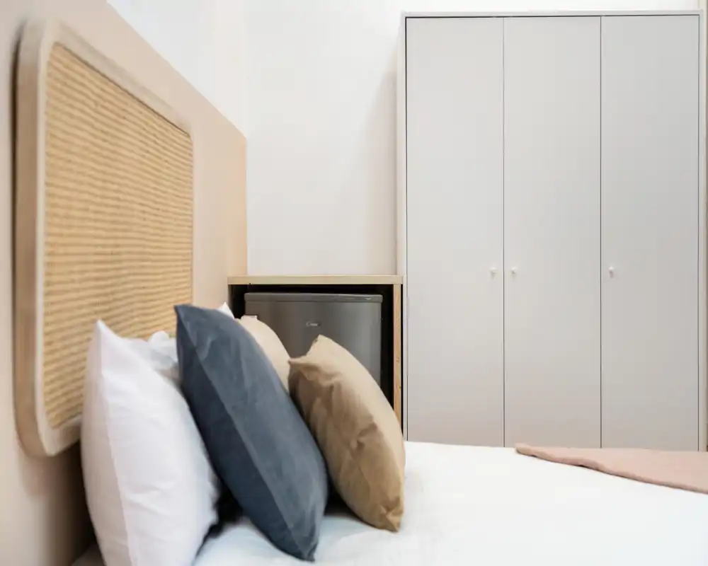 Photo lumineuse d'une chambre montrant un lit avec coussins superposés, une tête de lit tressée et une armoire en arrière-plan — propre, moderne et accueillante.