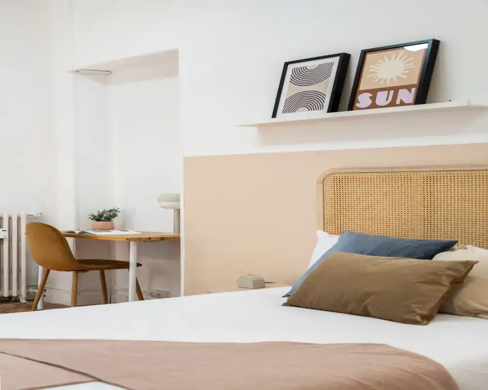 Chambre lumineuse et élégante avec une tête de lit en tressé, des coussins superposés et une palette de couleurs neutres. Un petit bureau et une chaise sont visibles en arrière-plan, pratique pour un peu de travail.