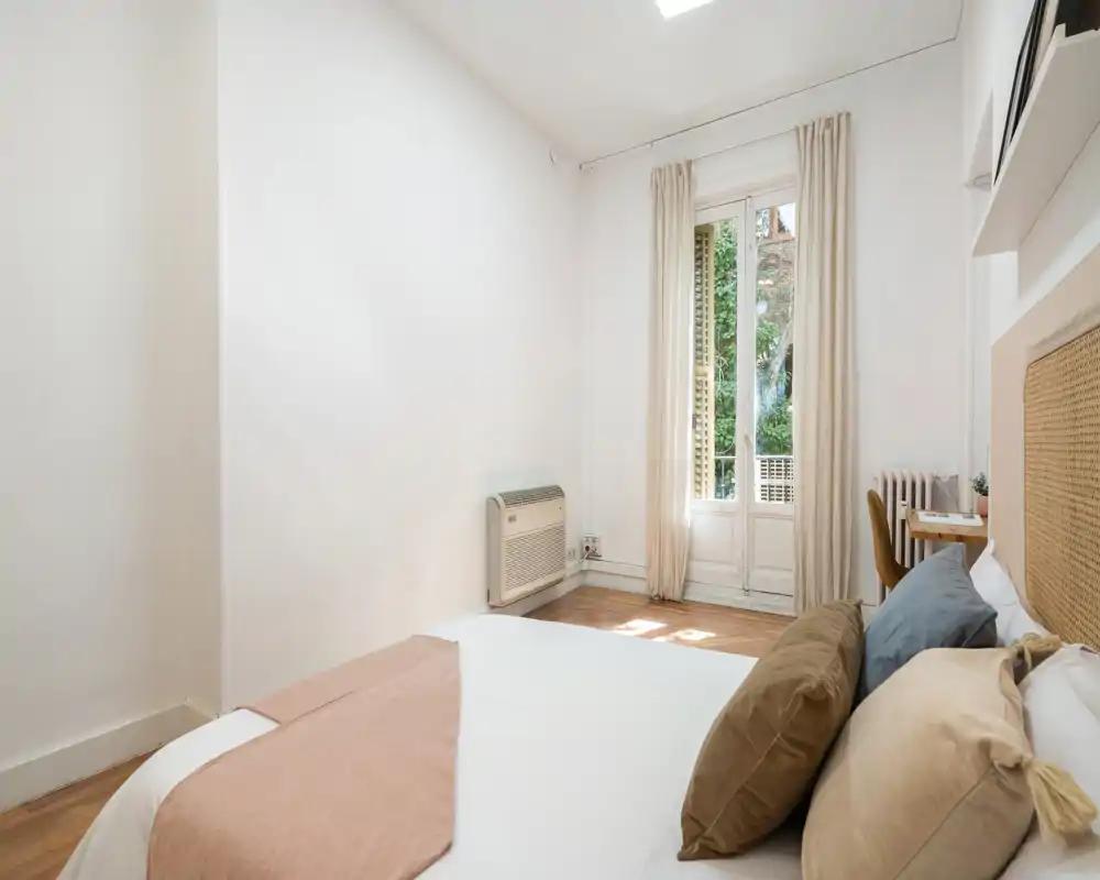Chambre lumineuse et aérée avec un lit bien fait, des coussins confortables et de la lumière naturelle provenant d'une porte-fenêtre donnant sur la verdure.