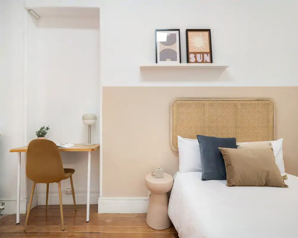 Chambre lumineuse et minimaliste avec une tête de lit en cannage, coussins superposés et une table de chevet soignée. Un petit bureau et une chaise à gauche forment un coin nuit/travail fonctionnel.