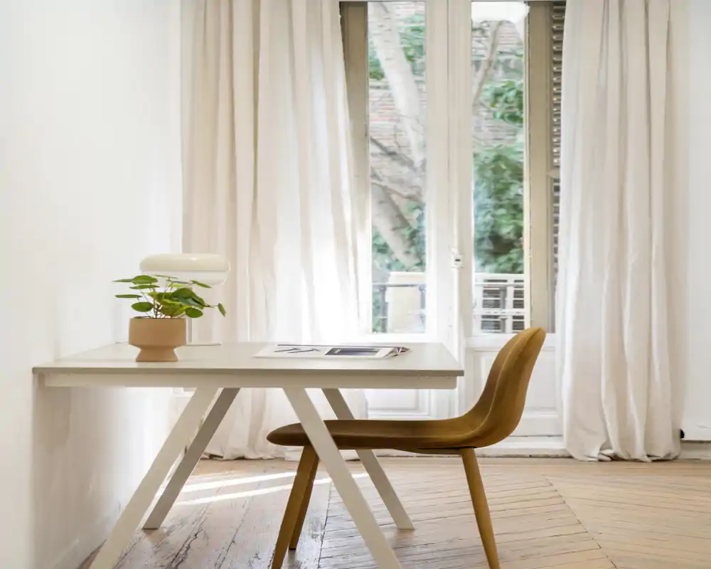Espace de vie lumineux et minimaliste avec un bureau simple, une plante et une chaise aux tons chaleureux face à de grandes fenêtres-fenêtres laissant entrer beaucoup de lumière naturelle.