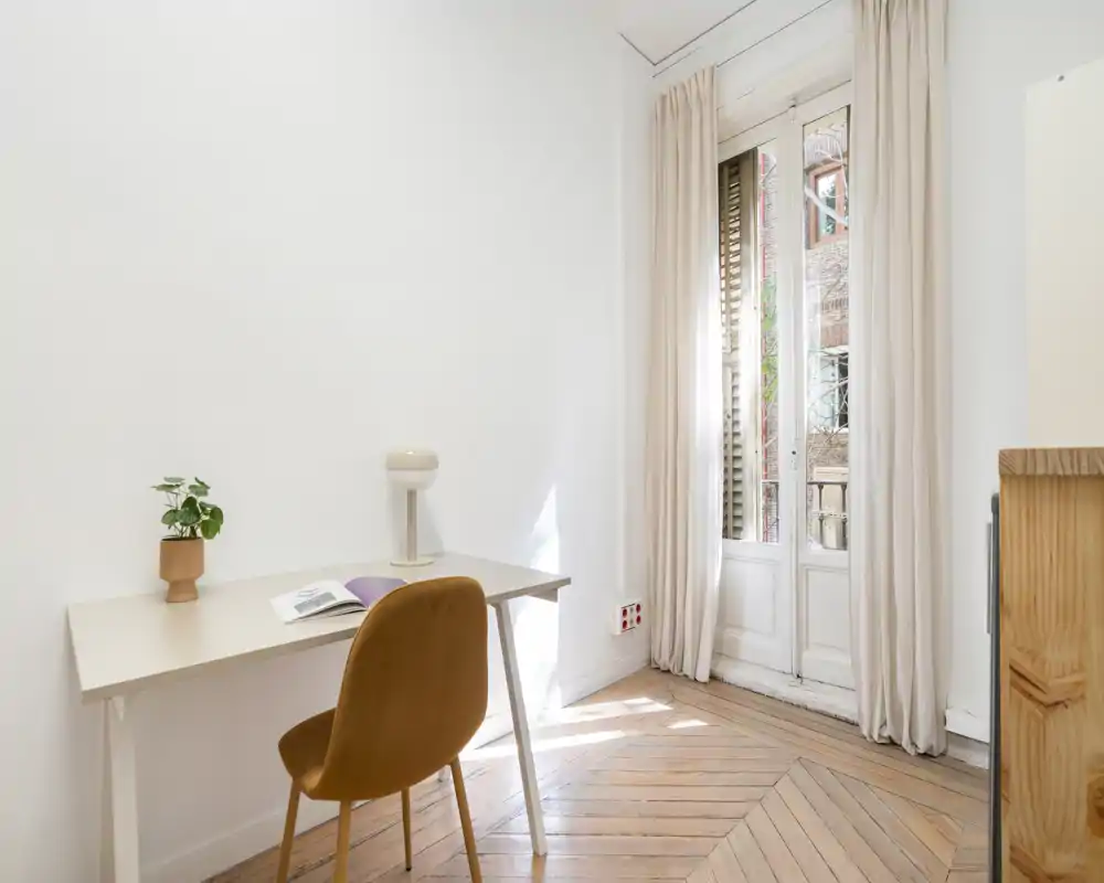 Pièce lumineuse et minimaliste avec un petit bureau et une chaise confortable à côté de grandes portes-fenêtres qui inondent l'espace de lumière naturelle — parfait pour un coin bureau.