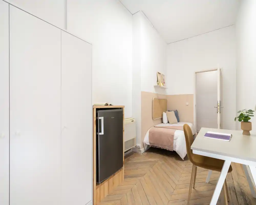 Chambre simple lumineuse et soignée avec un lit confortable, un petit bureau, une armoire et un parquet moderne — un espace chaleureux et bien éclairé, idéal pour une personne.