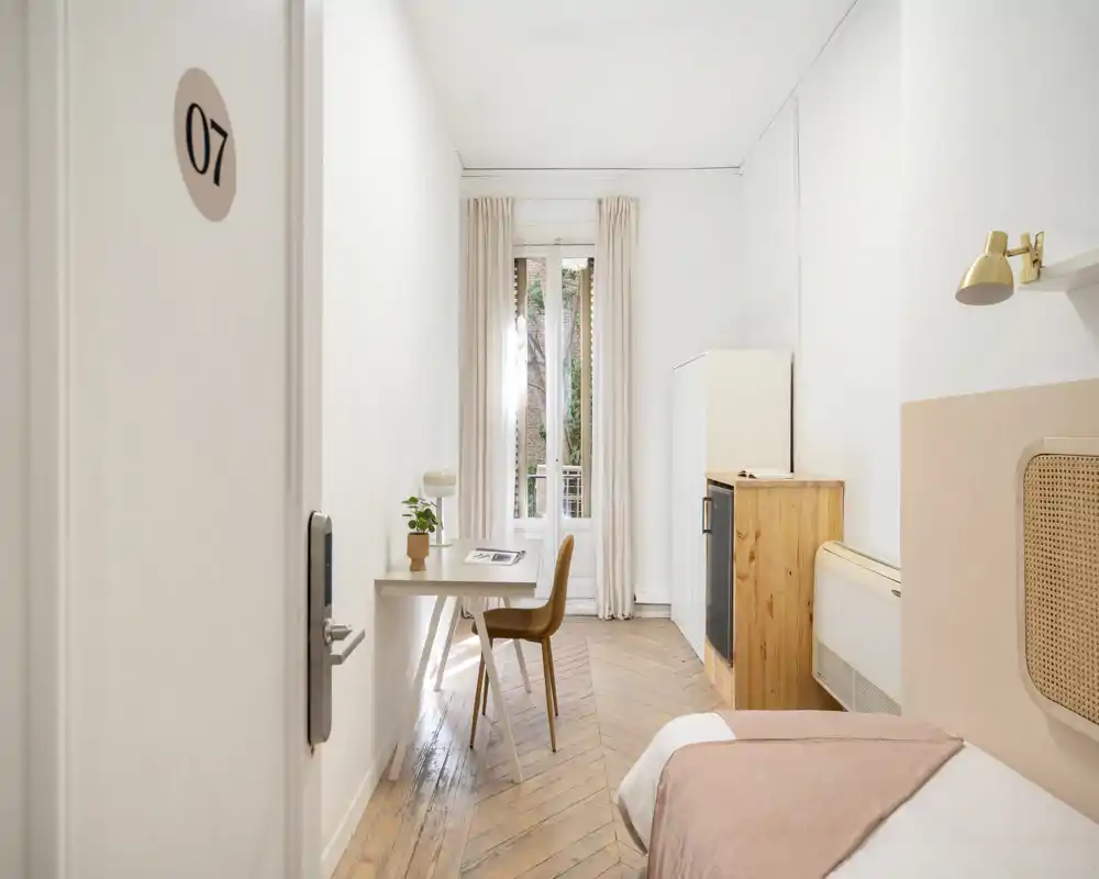 Chambre simple lumineuse et minimaliste avec lit soigné, parquet en bois, bureau près de la fenêtre et tons neutres — idéale pour un séjour confortable.