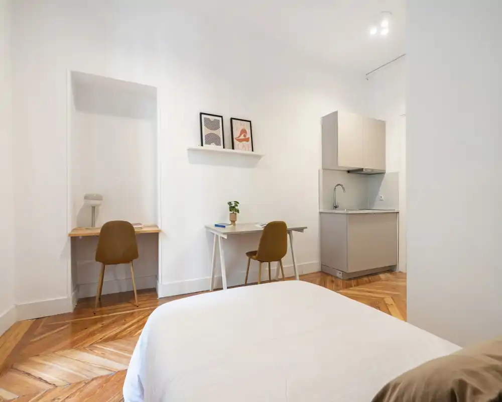 Chambre lumineuse et minimaliste avec un lit double au premier plan, deux bureaux avec chaises le long du mur et une kitchenette compacte. Décor épuré et parquet chaleureux pour un espace accueillant, parfait pour de courts séjours.