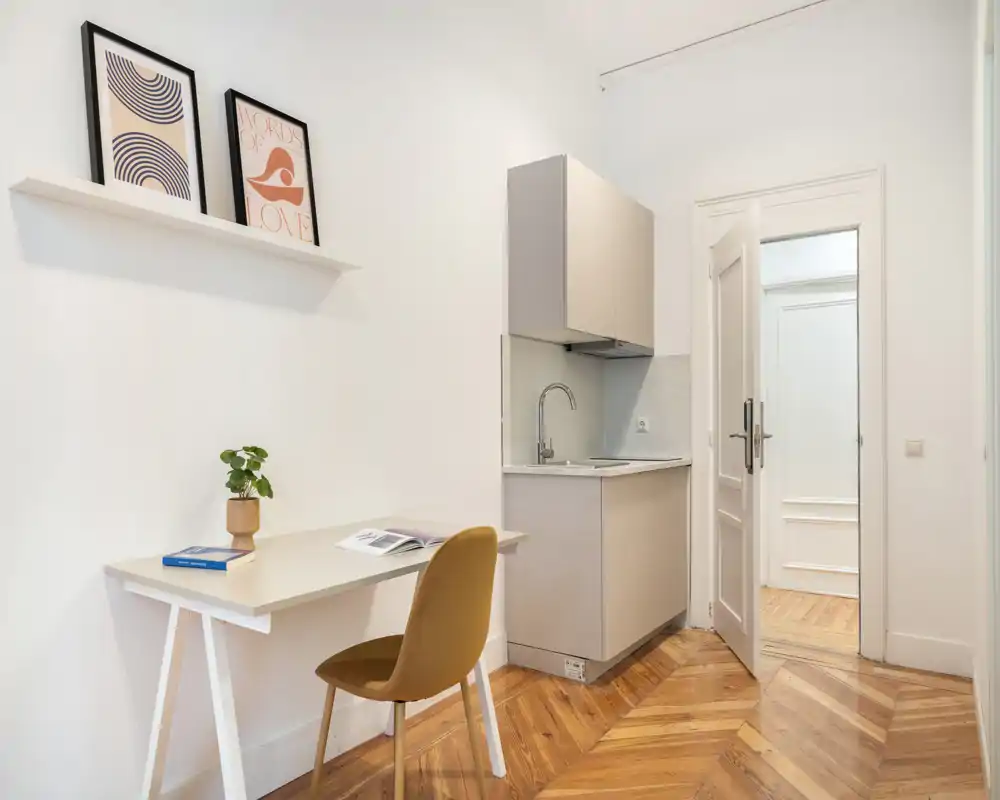 Coin cuisine compact avec petit évier, meubles minimalistes et un bureau; lumineux, soigné, avec un parquet en chevrons chaleureux et des cadres décoratifs.