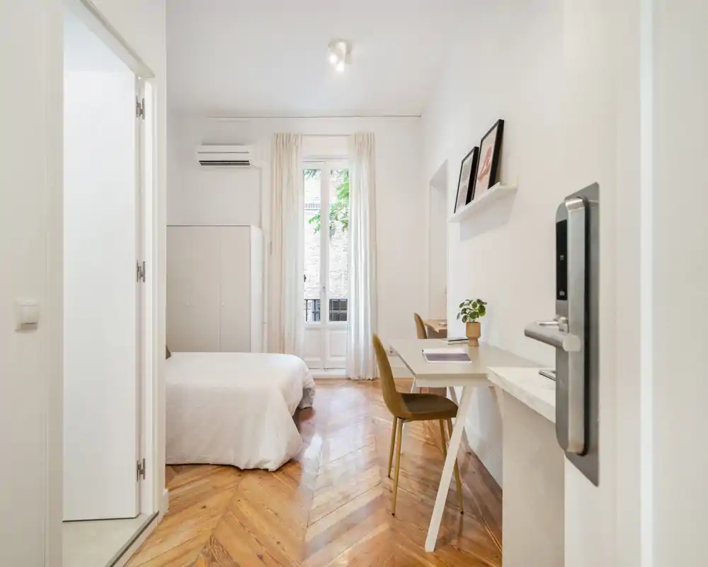 Chambre lumineuse et élégante avec un lit confortable, parquet en chevrons et un petit espace bureau près de la fenêtre. La lumière naturelle et la décoration neutre offrent une ambiance calme et accueillante.