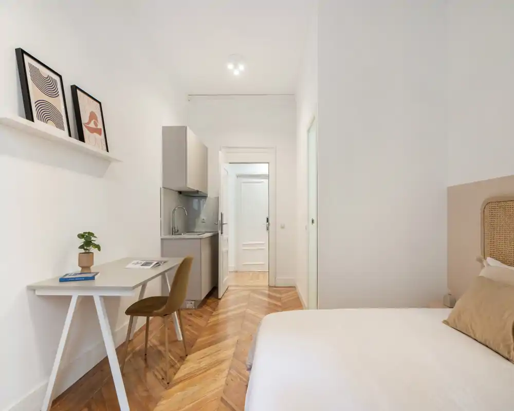 Chambre lumineuse et bien mise en scène avec lit visible, kitchenette compacte, espace bureau et parquet en chevrons élégant — idéale pour un studio.