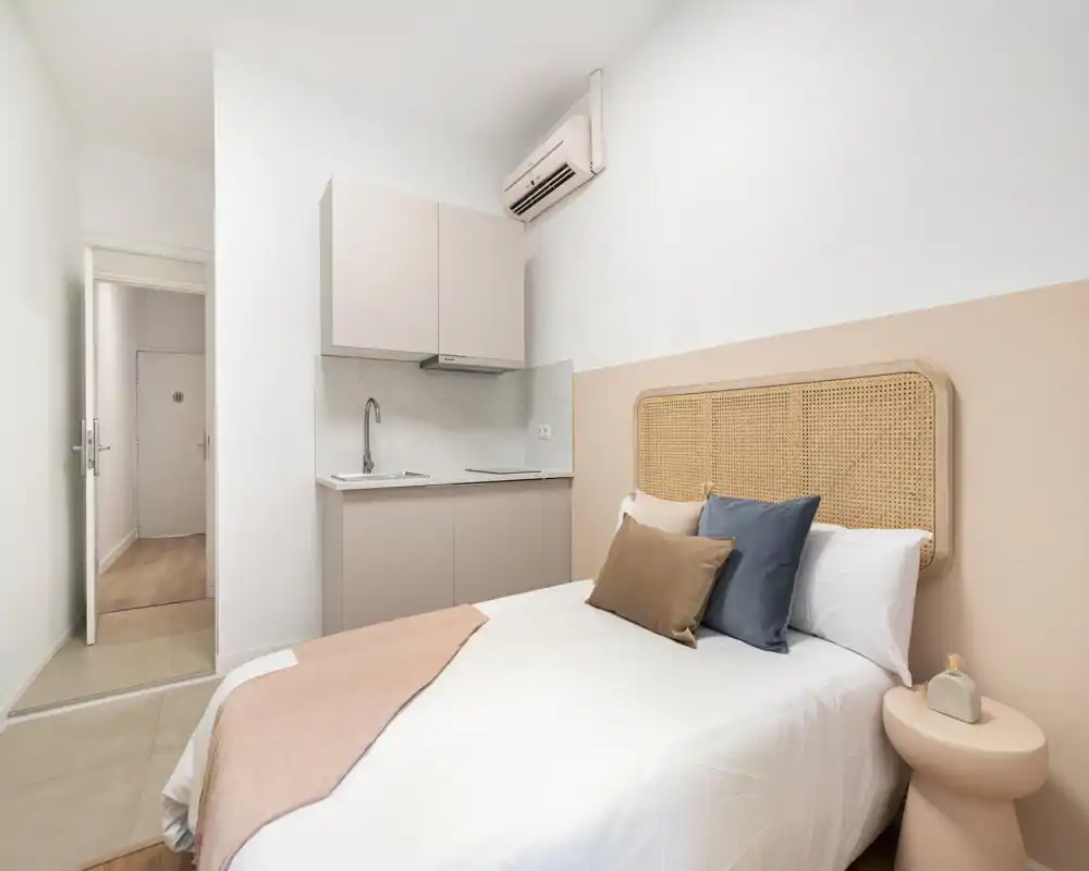Chambre lumineuse et bien mise en scène avec une tête de lit tressée et une kitchenette compacte contre un mur. Tons neutres, coussins et textiles doux pour une ambiance calme et accueillante idéale pour un studio.