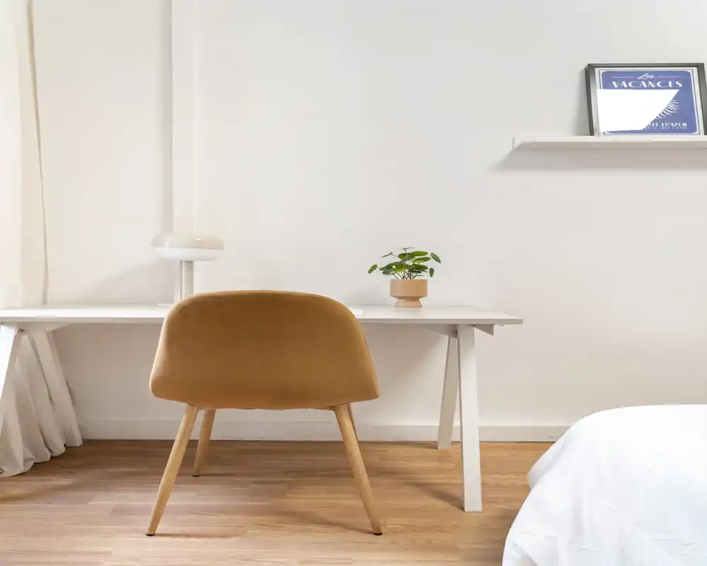Coin chambre lumineux et minimaliste avec un bureau blanc, une chaise rembourrée chaleureuse et une petite plante en pot. Une portion du lit est visible sur la droite, créant une ambiance calme et aérée, idéale pour une location compacte.