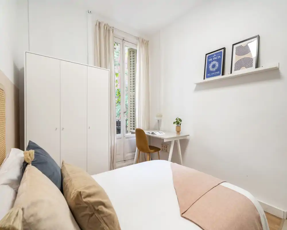Chambre lumineuse et au style doux avec un lit double confortable au premier plan, une armoire blanche et un petit bureau près de la fenêtre laissant entrer la lumière naturelle.