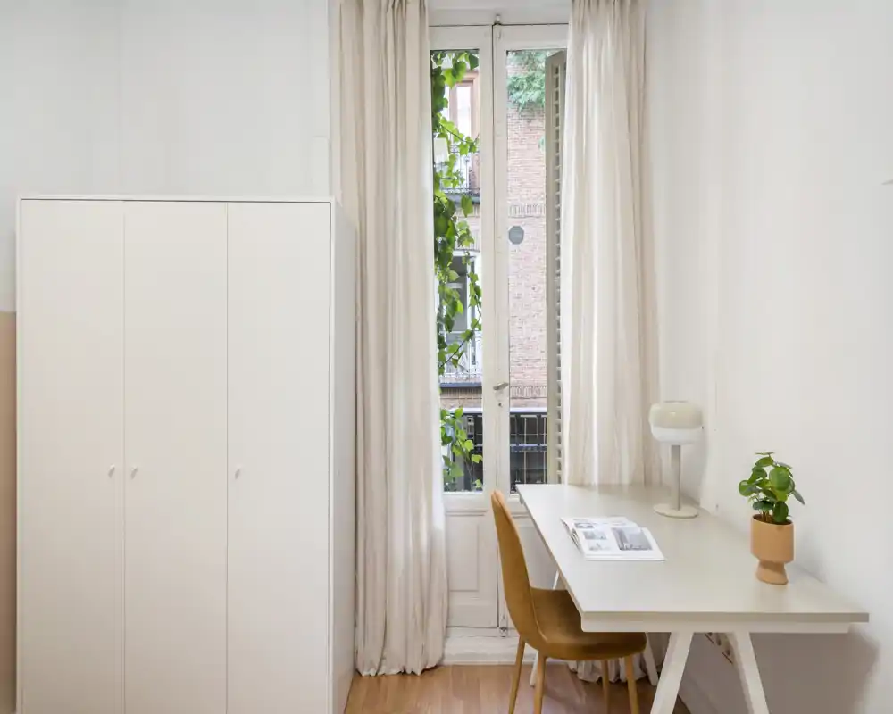 Coin de pièce lumineux et minimaliste avec une armoire blanche, un bureau simple et une chaise devant une grande fenêtre laissant entrer la lumière naturelle. Rideaux neutres et petite plante apportent une ambiance calme et fonctionnelle.