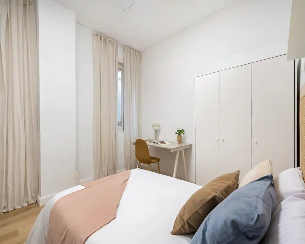 Dormitorio moderno y luminoso con cama bien vestida, textiles suaves y un pequeño escritorio junto a la ventana — ideal para descansar y trabajar ocasionalmente.