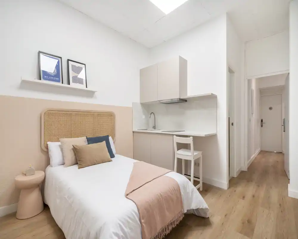 Estudio-dormitorio luminoso y moderno con cama doble, cabecero de mimbre y kitchenette compacta. Tonos neutros y textiles cálidos sobre suelo efecto madera que crean un ambiente acogedor, perfecto para alquileres de corta estancia.