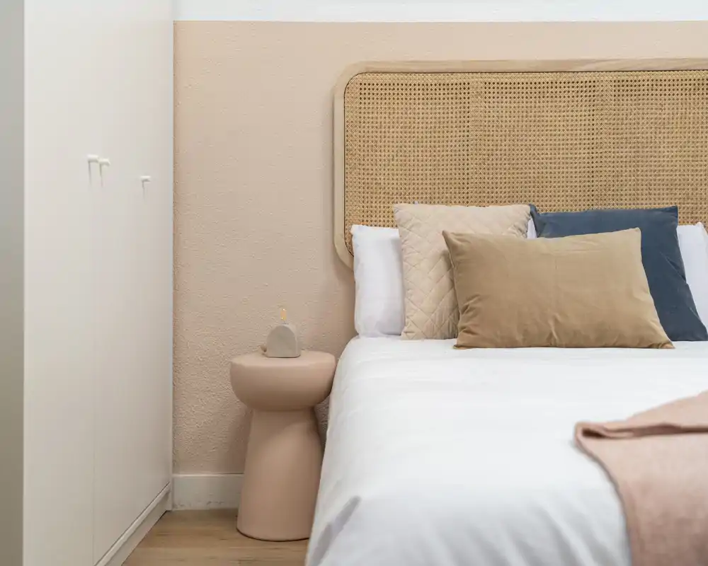 Rincón de dormitorio luminoso y estilizado con cabecero de rejilla, cojines decorativos y mesita suave — un espacio de descanso moderno y acogedor.