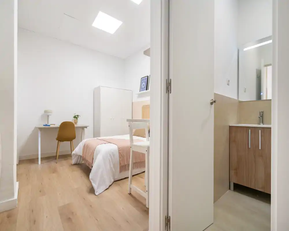 Dormitorio luminoso y minimalista con cama doble vestida en ropa blanca y un plaid rosa, un pequeño escritorio y silla, armario y suelos de madera cálida. La imagen muestra una zona de descanso limpia y bien iluminada con el baño en suite visible por la puerta abierta.