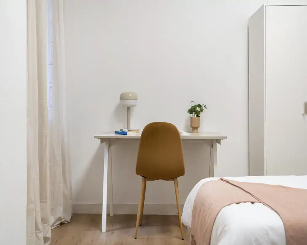 Rincón de dormitorio minimalista y luminoso con el borde de la cama visible, un escritorio sencillo con lámpara y una silla, y un armario — acogedor, moderno y ordenado.