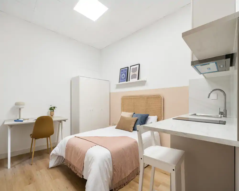 Dormitorio luminoso y bien arreglado con cama individual y cabecero de mimbre, escritorio y armario; se aprecia una pequeña cocina con fregadero y placa a la derecha, ideal para un estudio.