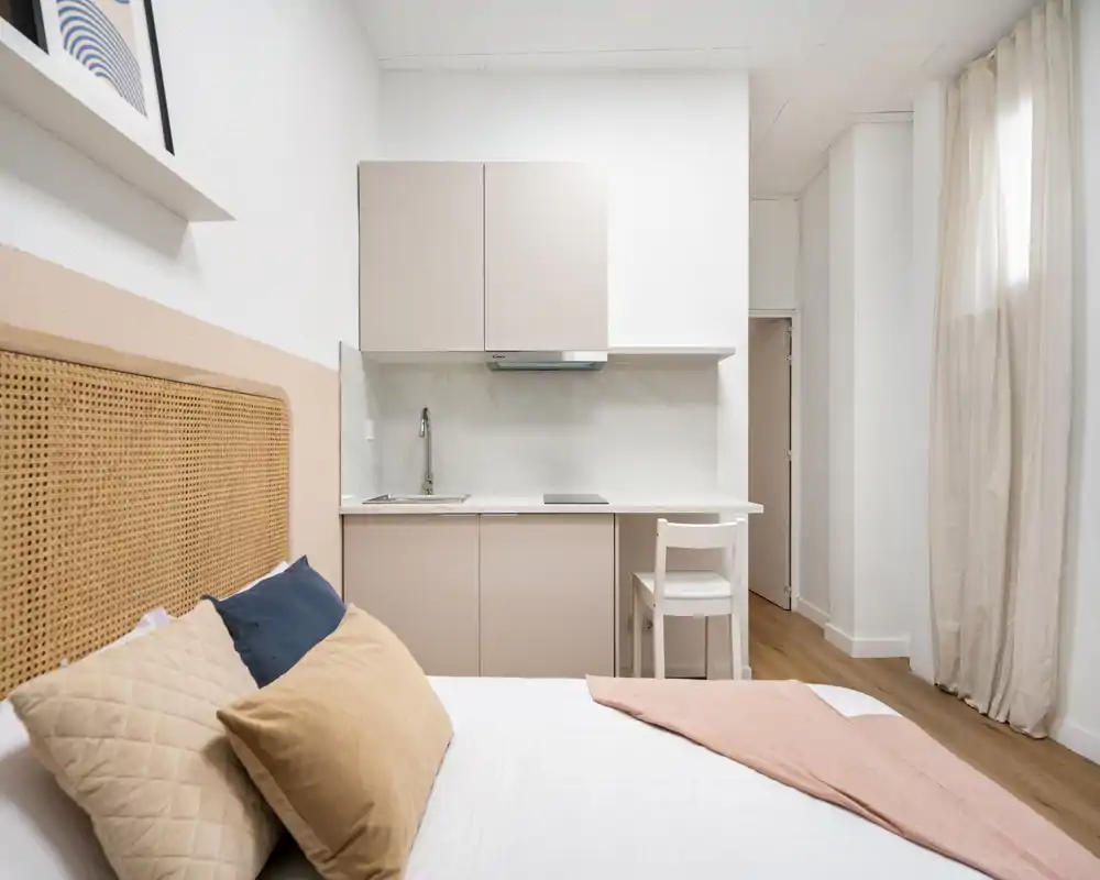 Estudio dormitorio luminoso y moderno con cama cómoda en primer plano y una cocina compacta al fondo; tonos neutros, textiles suaves y líneas limpias crean un ambiente acogedor y contemporáneo.