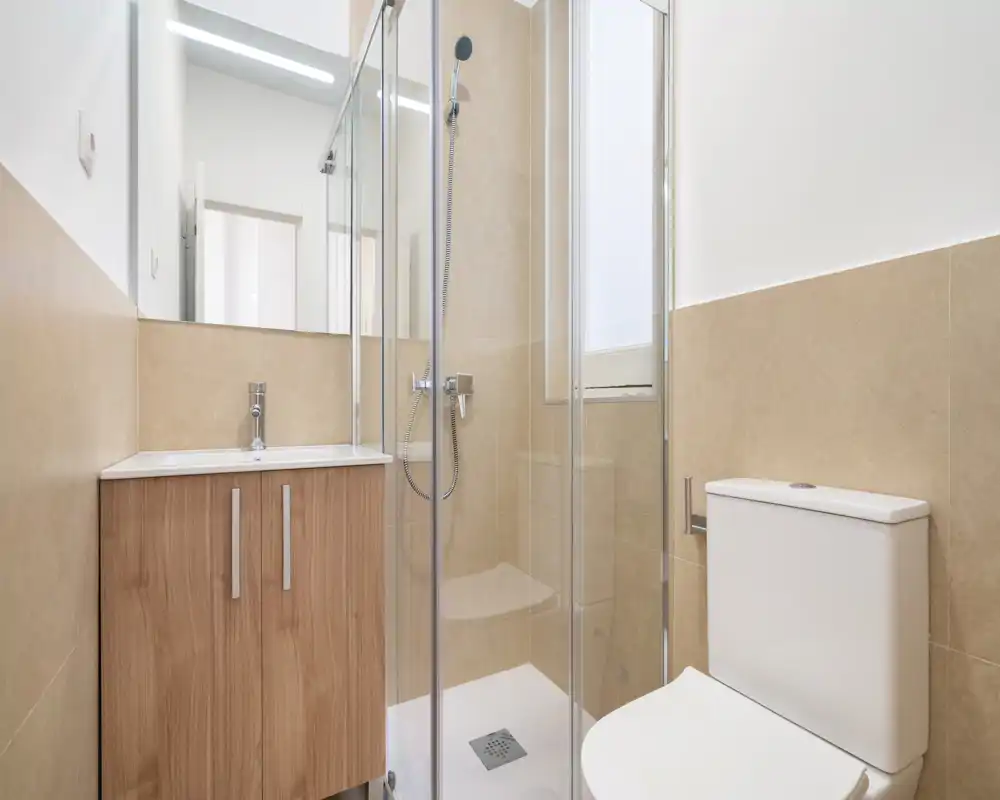 Baño moderno y compacto con ducha acristalada, alcachofa de pared, mueble de lavabo con efecto madera y inodoro compacto. Azulejos beige y espejo alto crean un espacio luminoso y minimalista.