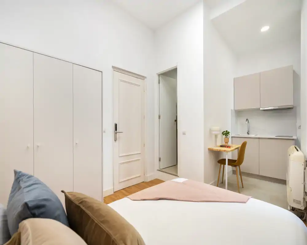 Dormitorio estudio moderno y luminoso con cama doble cómoda, armario empotrado y una pequeña cocina con mesa de comedor/trabajo, perfecto para alquiler tipo estudio.