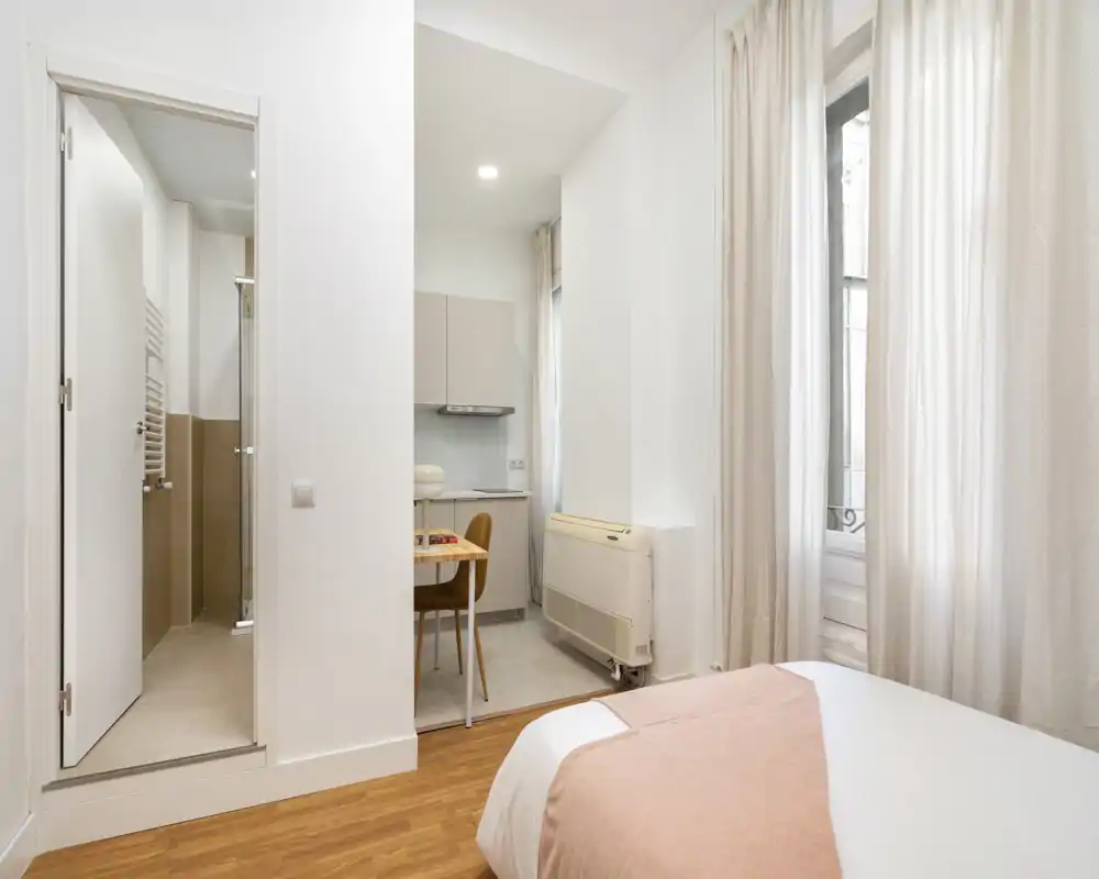Dormitorio compacto y luminoso con cama doble visible, textiles suaves y ventana que aporta luz natural; pequeña cocina y baño visibles a través de aberturas contiguas.