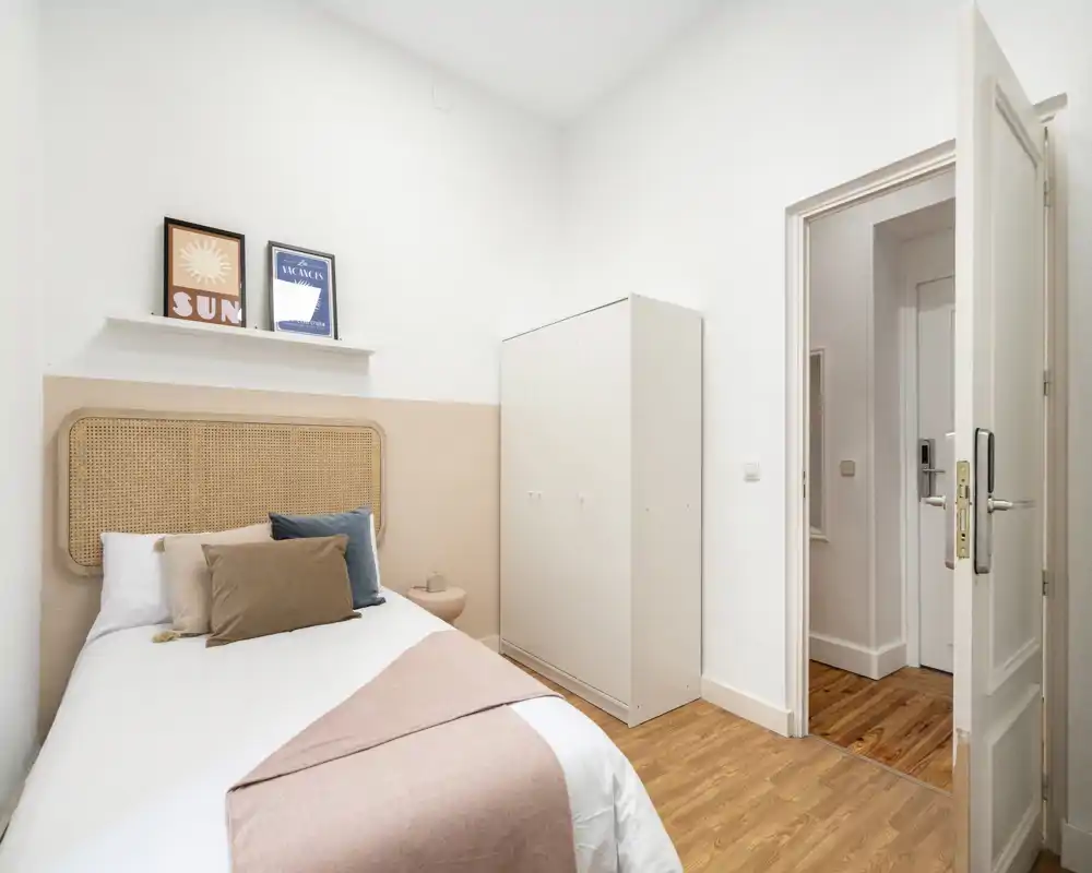 Dormitorio individual luminoso y bien decorado con cabecero de ratán, ropa de cama suave y cojines coordinados; armario blanco y suelo de madera para un ambiente acogedor y minimalista.