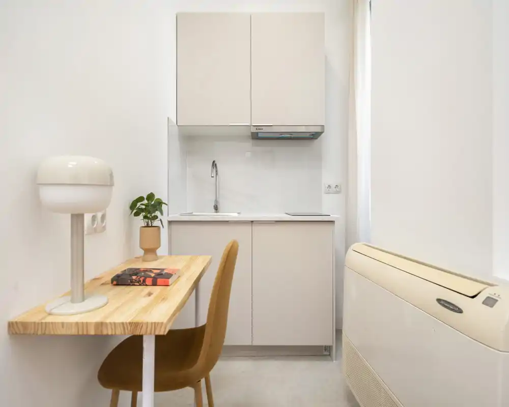 Kitchenette compacta y moderna con armarios blancos, fregadero y una pequeña mesa de madera con silla — ideal para un estudio.