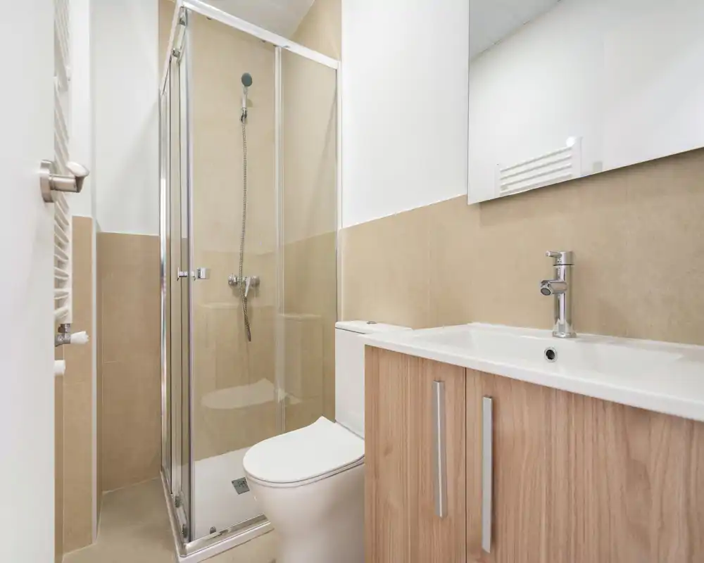 Baño moderno y luminoso con mampara de ducha de vidrio, inodoro y mueble lavabo; azulejos beige y líneas limpias para un aspecto minimalista.