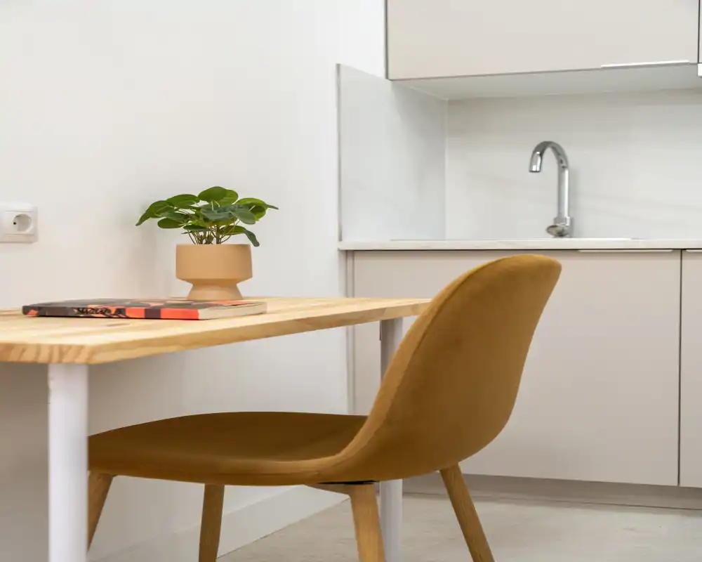 Rincón de cocina moderno y compacto con mesa de madera, silla tapizada y área de fregadero minimalista. Una pequeña planta y un libro añaden calidez.
