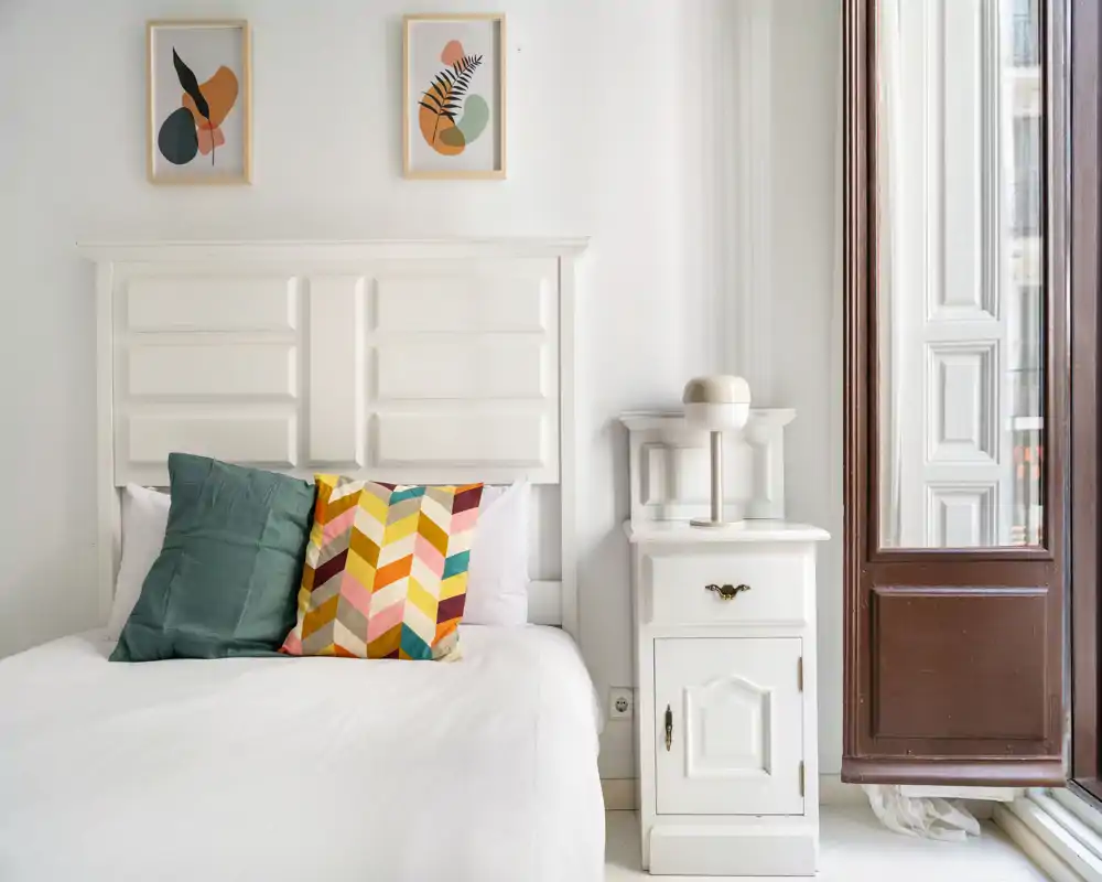 Camera da letto luminosa e curata con testata bianca, biancheria fresca e cuscini colorati accanto a un comodino e a una grande finestra che illumina la stanza.