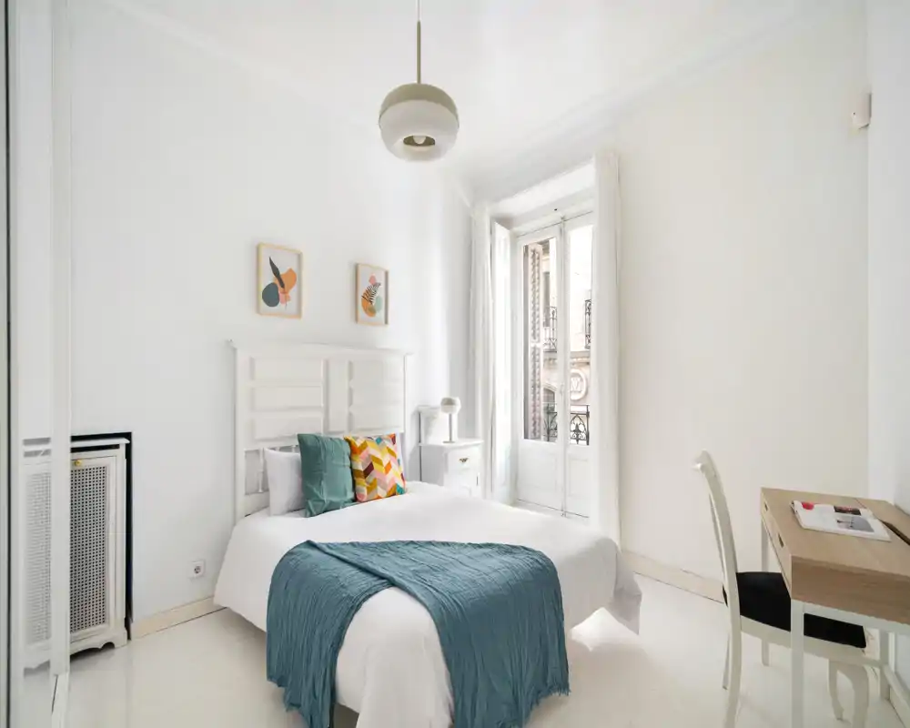 Camera da letto luminosa e minimal con letto matrimoniale ben rifatto, cuscini decorativi e un plaid turchese; grande finestra con luce naturale e una scrivania piccola.