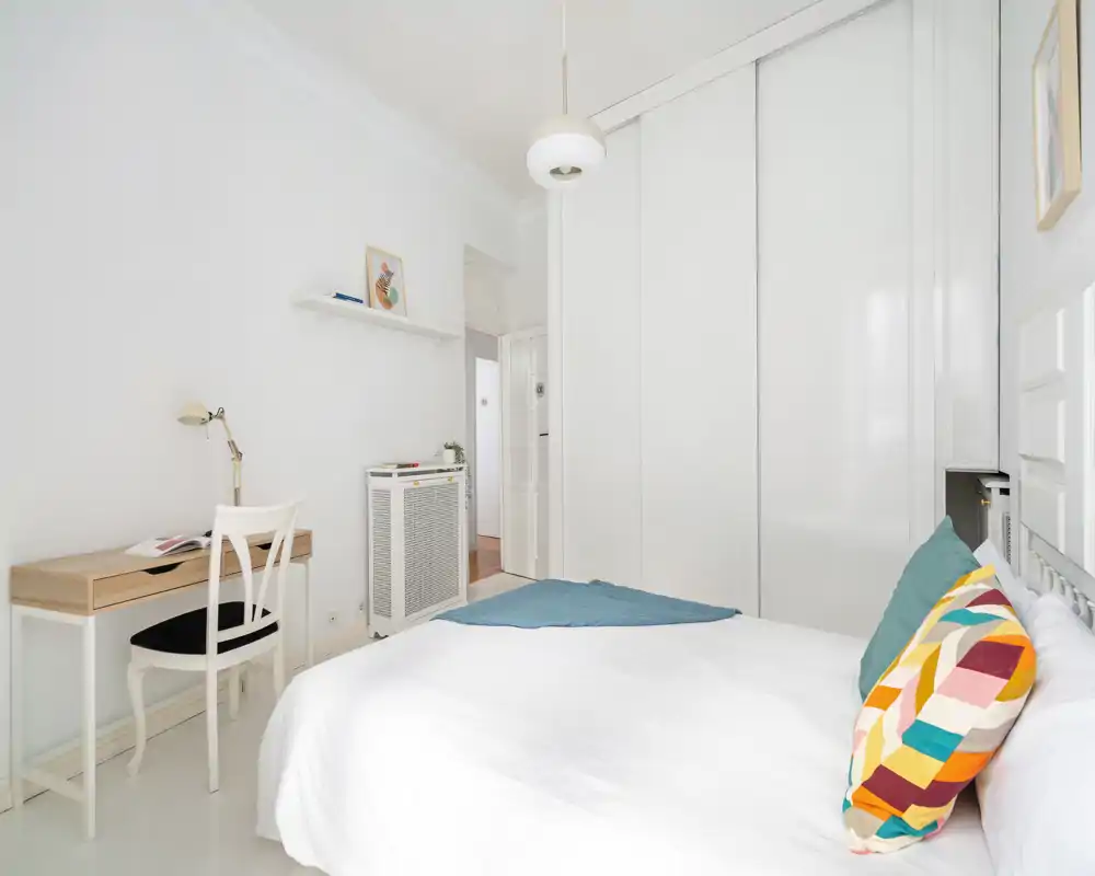 Camera da letto luminosa e minimal con letto bianco, cuscino colorato e scrivania semplice: ideale per un annuncio moderno e pulito.