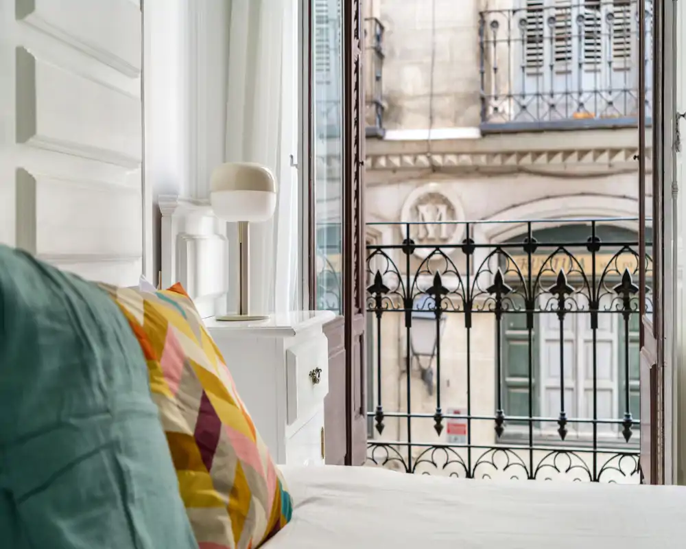 Angolo camera da letto luminoso e di stile con cuscini colorati, lampada da comodino e balcone aperto con vista sulla strada — ambiente accogliente e ben illuminato, ideale per un soggiorno in città.