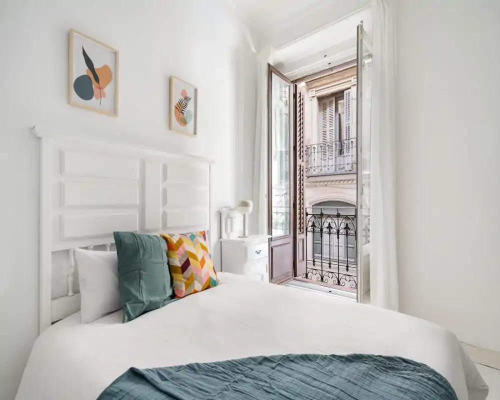 Camera da letto luminosa e ariosa con testata bianca, cuscini colorati e una finestra in stile francese aperta che porta luce naturale e vista sulla facciata esterna.