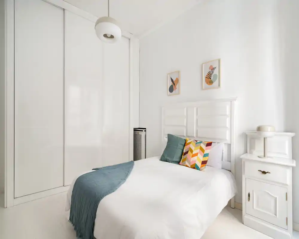 Camera da letto luminosa e minimal con testiera bianca, cuscini colorati e un plaid turchese. Ampio armadio a muro e comodino completano lo spazio ordinato e arioso, perfetto per annunci.