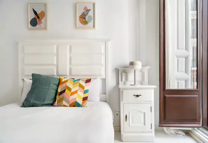 Dormitorio luminoso y con estilo con cabecero blanco, cojines decorativos de colores y mesita de noche junto a una gran ventana. La estancia resulta luminosa y acogedora, ideal para fotos de alquiler.
