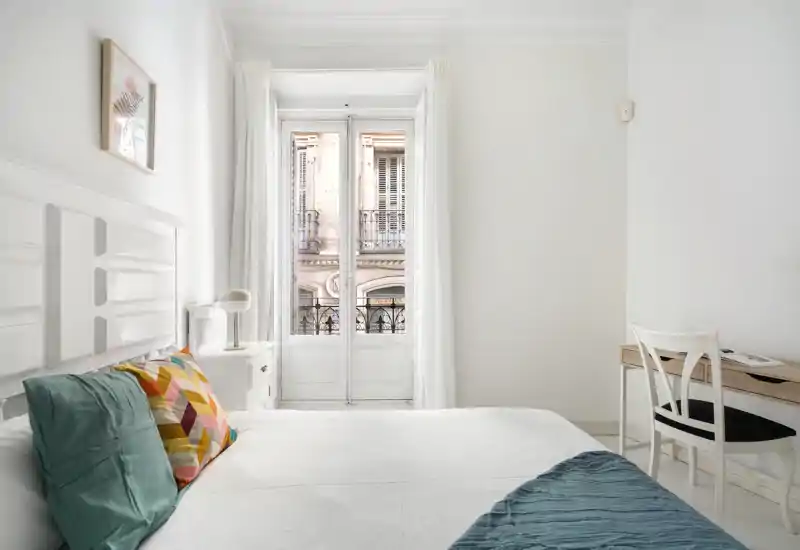 Camera da letto luminosa e minimal con ampio letto bianco, cuscini colorati, comodino e scrivania vicino a una finestra alta che illumina l’ambiente con luce naturale.