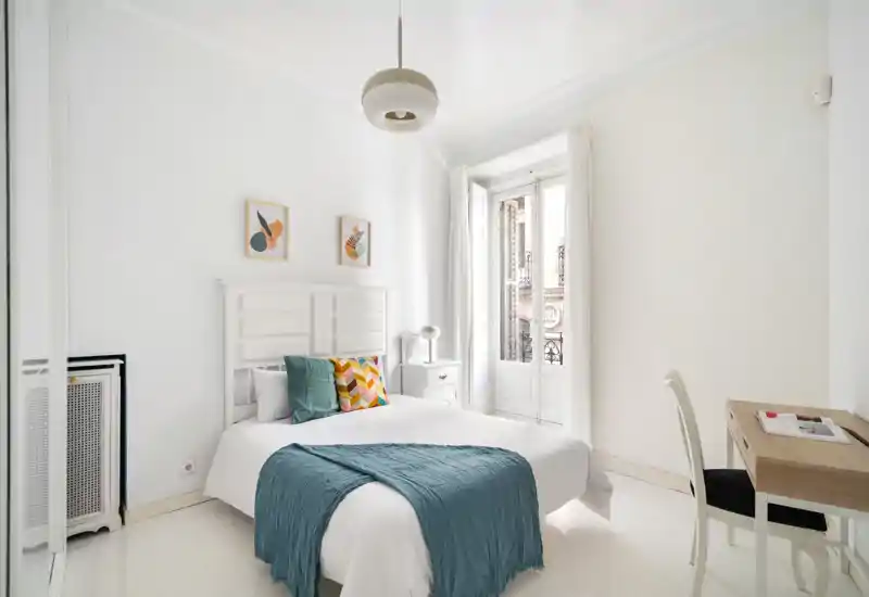 Dormitorio luminoso y aireado con cabecero blanco, cama doble bien hecha, cojines coloridos y plaid turquesa, escritorio y ventana alta que aporta luz natural.