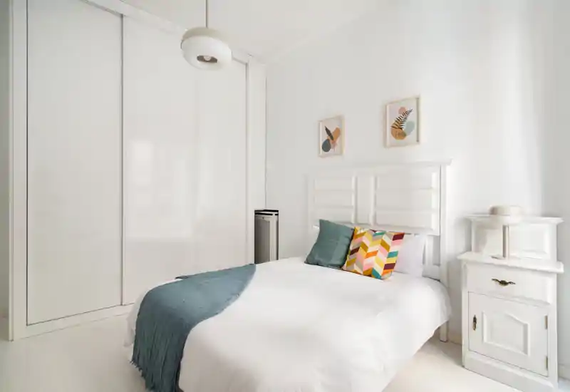Dormitorio luminoso y minimalista con cama doble centrada, cojines coloridos y una manta; gran armario empotrado y mesita moderna completan el espacio.
