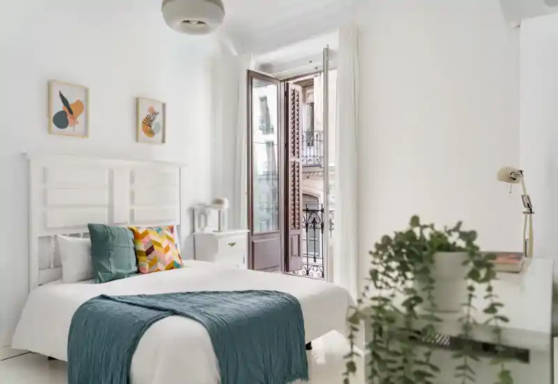 Dormitorio luminoso y aireado con cabecero blanco, ropa de cama en capas y una manta turquesa; puertas balconeras abiertas dejan entrar abundante luz natural.
