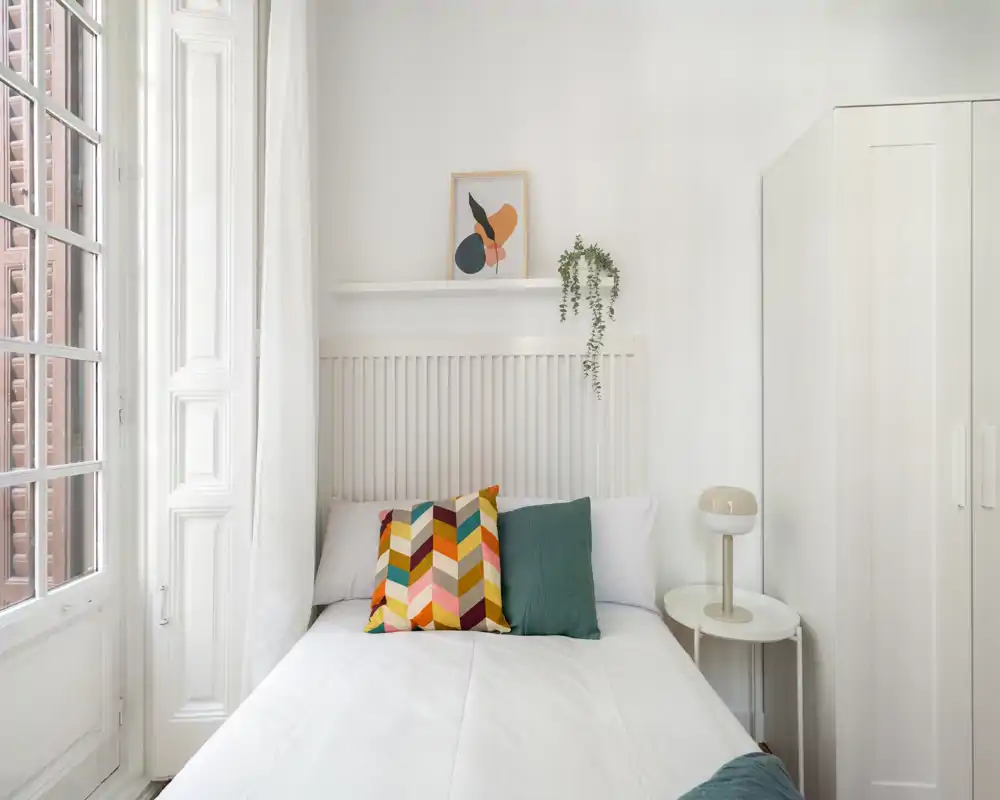 Chambre lumineuse et stylée avec un lit bien fait, coussins décoratifs colorés, table de chevet avec lampe et grand placard blanc — espace de nuit calme et aéré, éclairé par une large fenêtre.