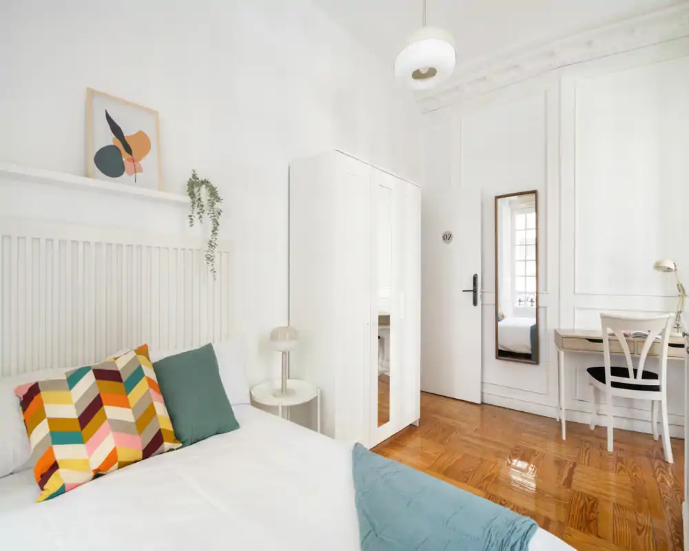 Chambre lumineuse et élégante aux murs blancs, parquet en bois, armoire et bureau. Le lit est préparé avec des coussins colorés et un plaid douillet, idéale pour des annonces mettant en avant confort et luminosité.